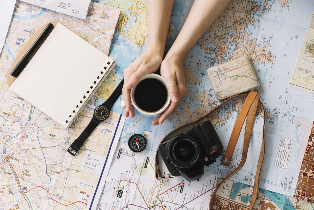 Mãos segurando copo de café com um caderno, máquina fotográfica, mapas e relógio do lado.