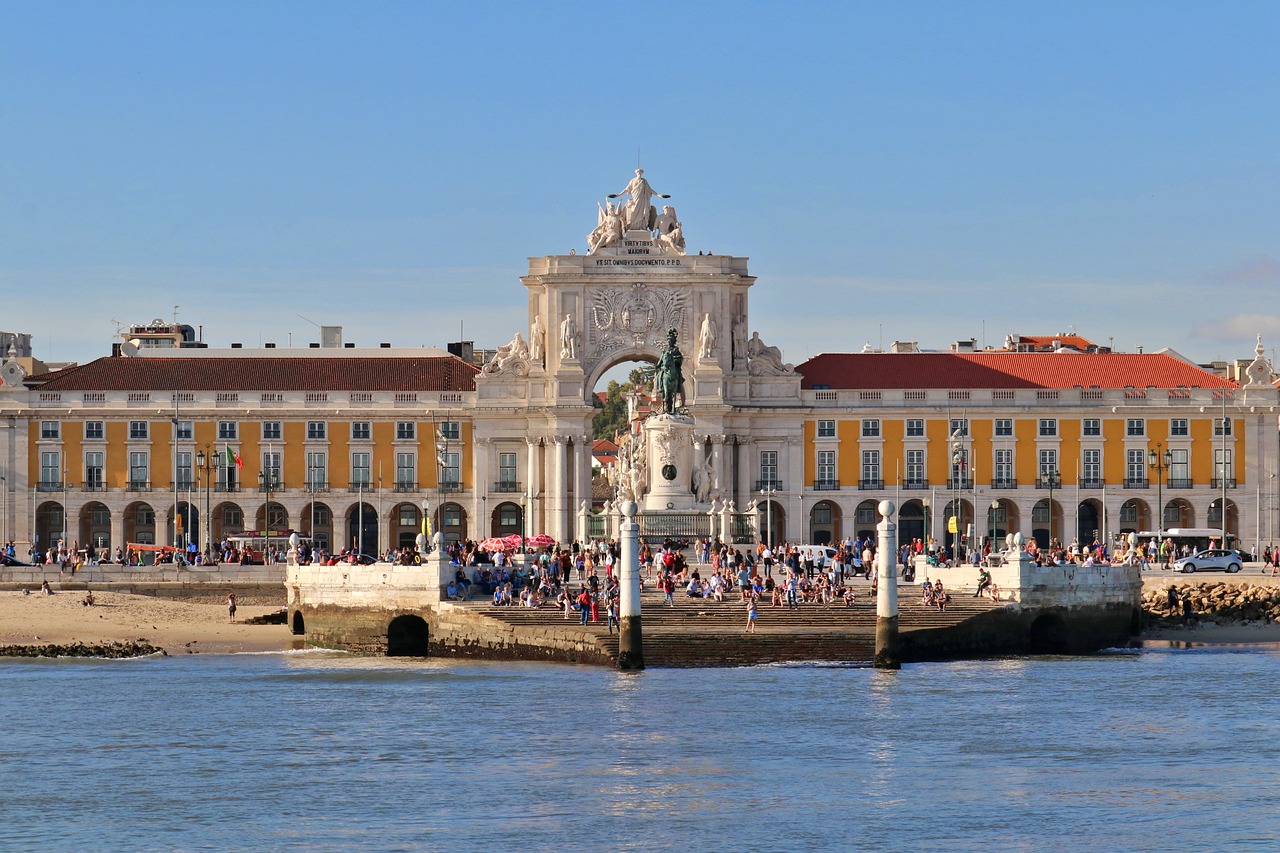 Praça do Comércio, torres do cais e estátua ao fundo. Pessoas sentadas na margem do rio Tejo. Lisboa, Portugal