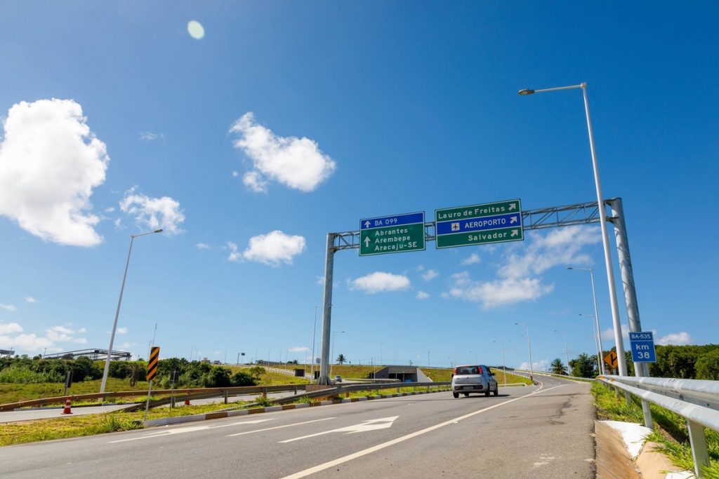 Imagem mostra a BR BA-093 que liga Salvador a demais cidades da região, possui um carro em movimento na estrada. 