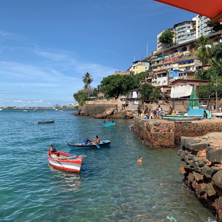 Praia da Gamboa em Salvador, Bahia - Na imagem mostra dois barcos no mar com pescadores em cima, atrás possui casas posicionadas sobre um morro.