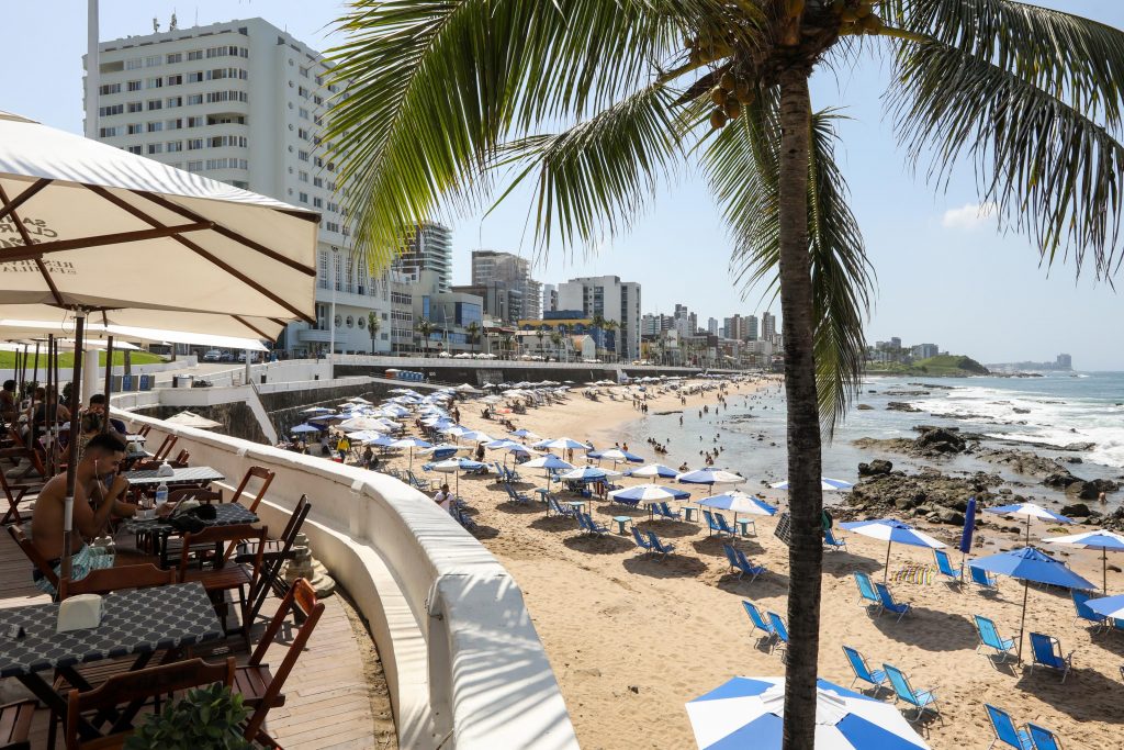 Praia do Farol da Barra - Salvador, Bahia - Na imagem mostra guarda-sóis na areia da praia com prédios posicionados atrás da avenida principal. 