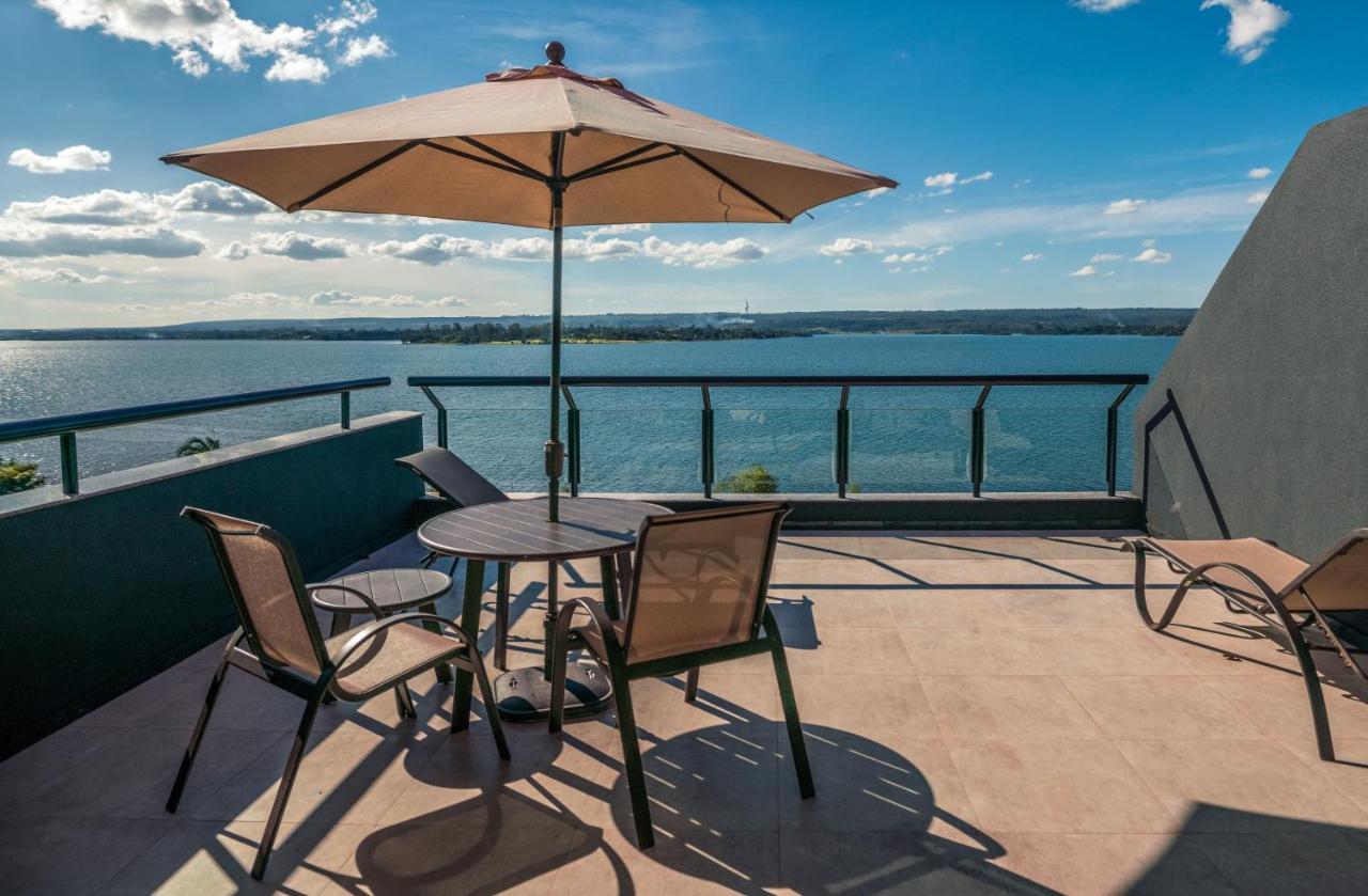 Varanda com mesa, guarda-sol e cadeiras. Lago Paranoá ao fundo. Royal Tulip Brasília Alvorada.