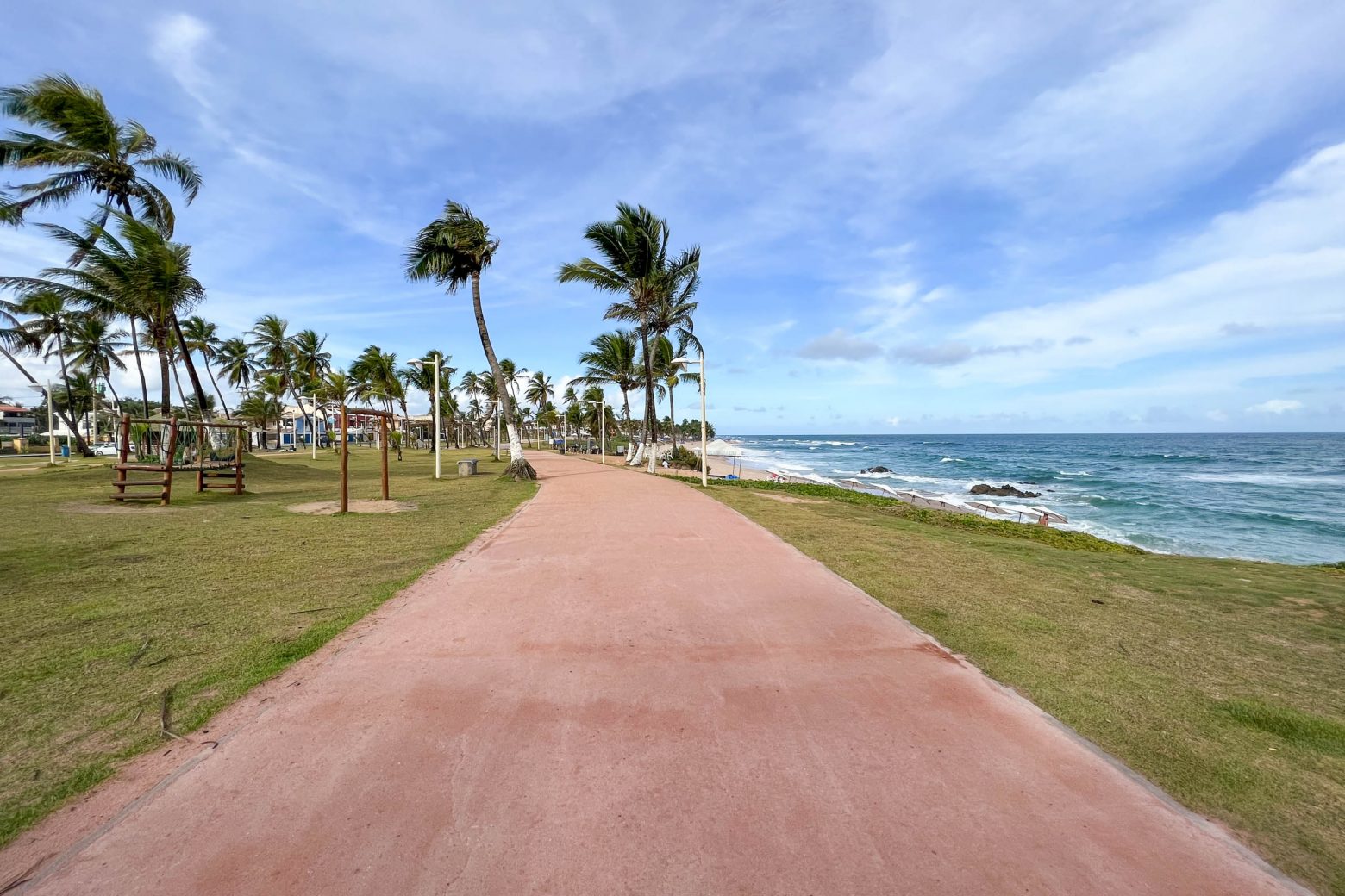 Praia de Stella Maris em Salvador, BA - Na imagem mostra o mar no canto lateral direito, e a frente uma calçada pintada de vermelho, com coqueiros a frente.