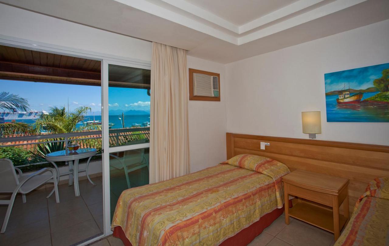 Hotel Atlântico Búzios - Quarto com cama, mesa lateral, abaju e varanda com vista para o mar.