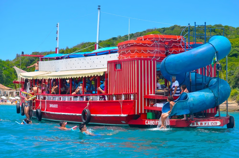 Escuna vermelha com toboágua e pessoas nadando no mar. Búzios.