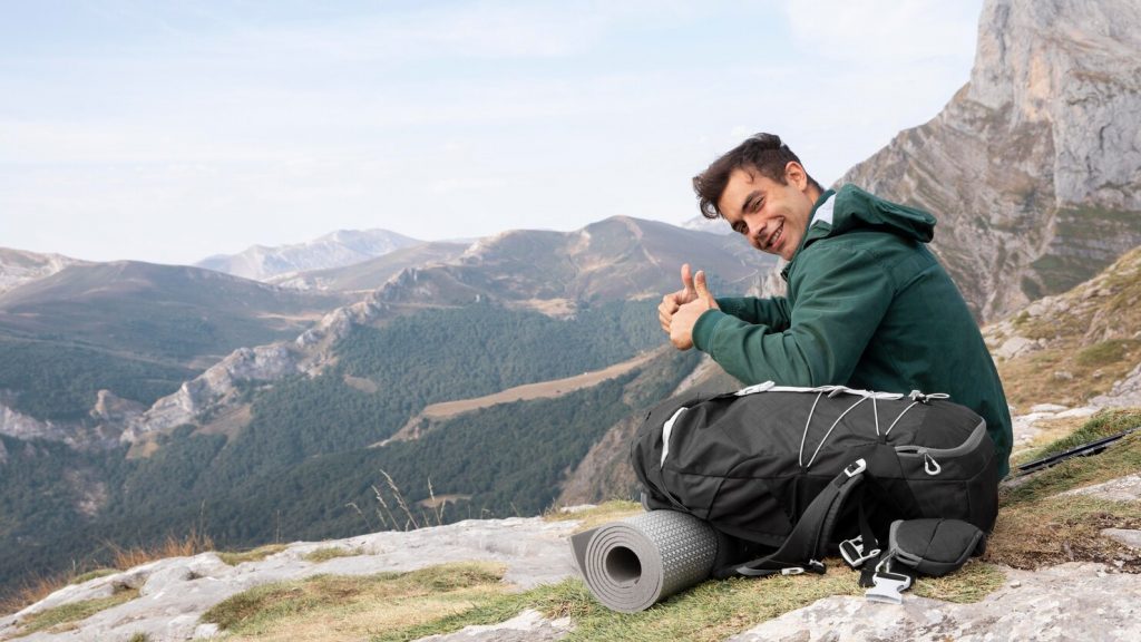 jovem sentado de frente para as montanhas. Tem mochila e tapete.